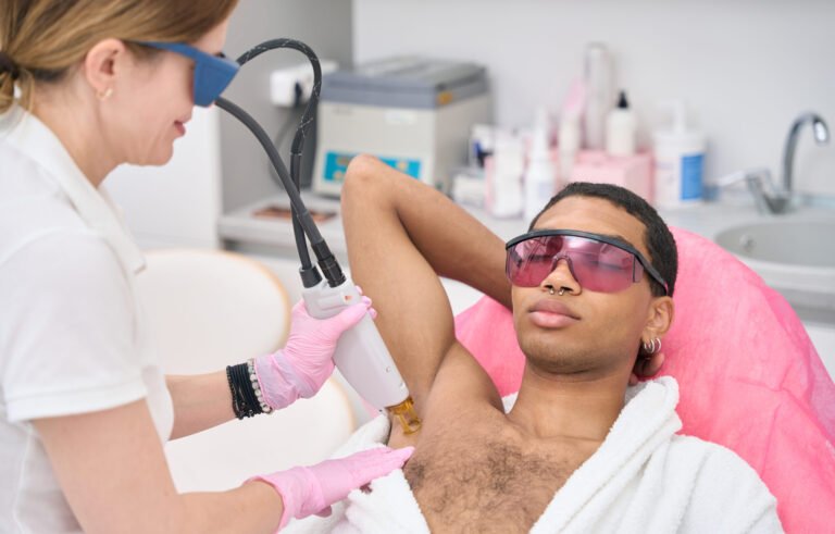 Beautician in nitrile gloves and safety goggles removing unwanted hair from man armpit using modern equipment