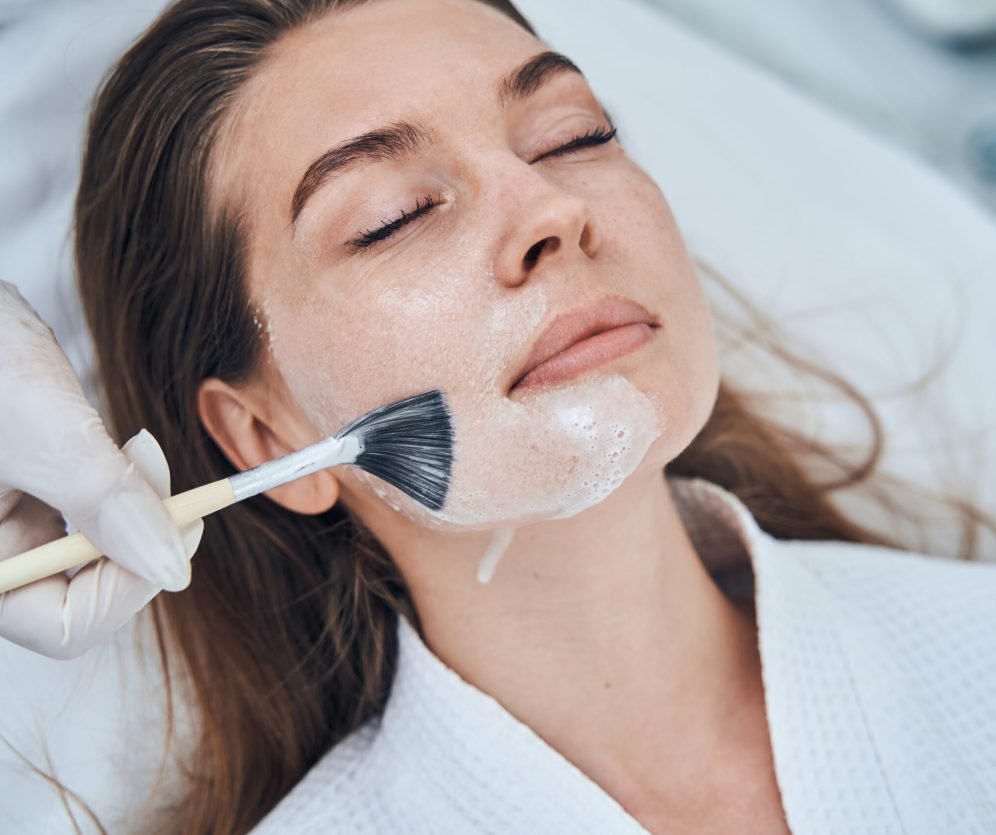 Competent cosmetologist wearing rubber gloves while using brush for applying mask on face