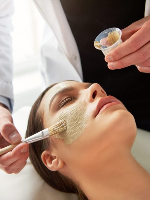 Male cosmetologist putting mask on young female face with a brush during cosmetic procedure.
